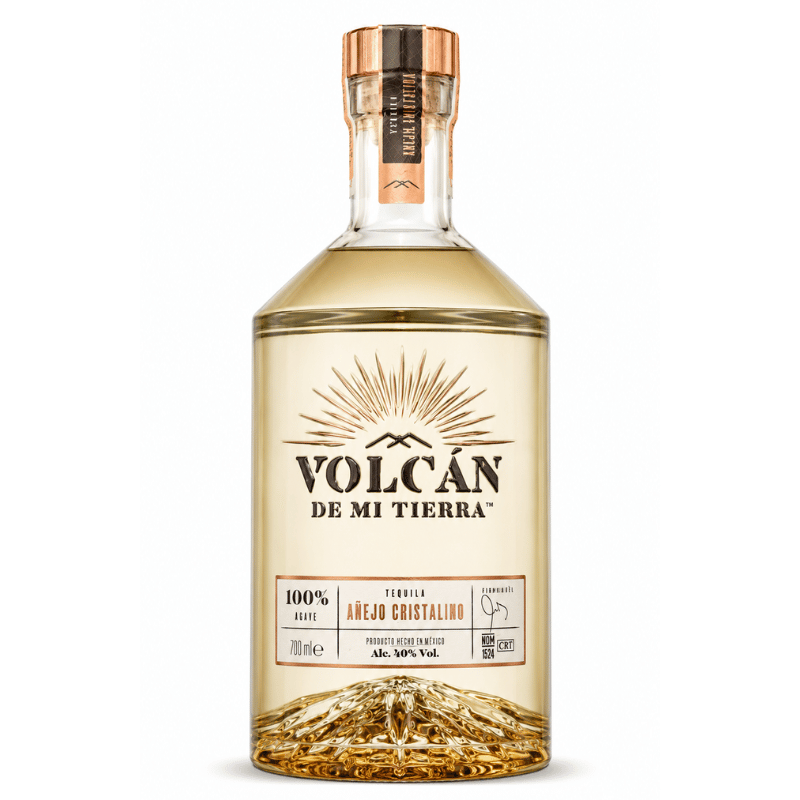 A bottle of Volcán de Mi Tierra tequila on a clean white background. The clear glass bottle shows the golden-amber liquid inside, featuring a metallic copper cap and a detailed label with a gold sunburst design and brown text.