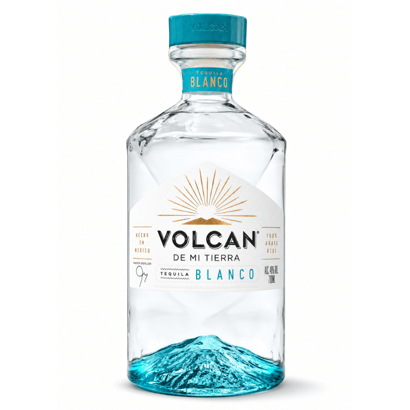 A bottle of Volcan De Mi Tierra Tequila Blanco on a clean white background. The clear glass bottle features a white label with a gold sun graphic and a bright blue mountain design at the bottom, topped with a teal cap.