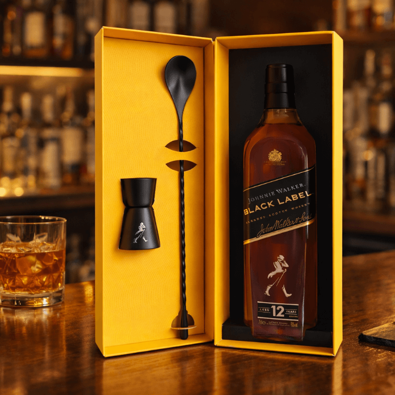 Johnnie Walker Black Label whisky bottle with bar tools in a yellow box on a wooden surface.